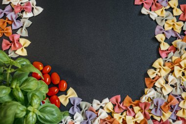 Renkli farfalle makarnası, kiraz domatesleri ve fesleğen yaprakları koyu dokulu arka planda bir çerçeve olarak dizilmiş. Kopya alanı olan İtalyan mutfağı kavramı.