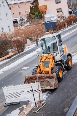 Sarı tekerlek yükleyici, kaldırılmış kovayla karları temizliyor ve kış yolu inşaatı sırasında bir yerleşim bölgesinin enkazını temizliyor. Önünde güvenlik bariyeri var..