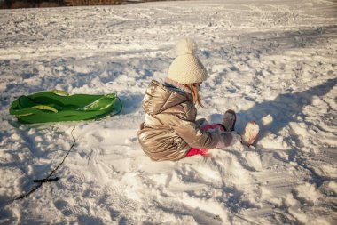 Küçük kız kış yürüyüşü sırasında yeşil kızağın yanında karlı zeminde oturuyor. Kış oyunu, çocukluk ve açık hava eğlence konsepti.