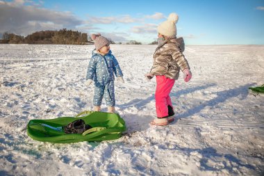 İki küçük kız kışın yeşil kızakla karlı bir tarlada birlikte oynuyorlar. Çocukluk, arkadaşlık, kış açık hava aktiviteleri ve aile eğlencesi konsepti..