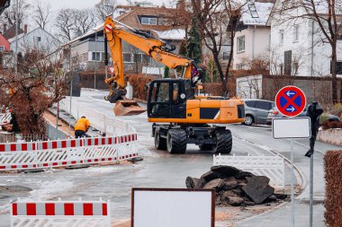 Sarı tekerlekli kazıcı ile inşaat işçileri kışın, kırmızı ve beyaz güvenlik bariyerleri, trafik işaretleri, toprak yığınları ve konut binalarıyla çevrili karlı bir şehir caddesini onarıyor.