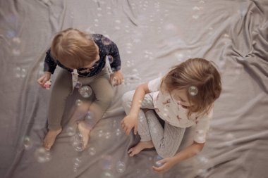 İki küçük kız kardeş birlikte yatakta oturuyor ve evlerinde sabun köpükleriyle oynuyorlar. Kardeş sevgisi, mutlu çocukluk ve aile bağı kavramı.