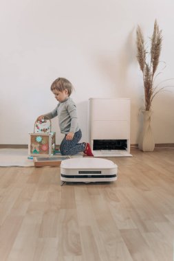 Arka planda küçük bir kızın bulanık bir silüetiyle, kaplamalı bir zeminde kuru temizleme yapan modern, beyaz bir robot elektrikli süpürgenin yakın çekimi..