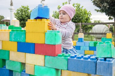 A girl standing and carefully placing a large blue toy block on top of a colorful wall outdoors.