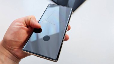 Modern bir akıllı telefonun, parlak beyaz bir arkaplanda insan eliyle tutulan kırık bir cam ekranı, cihaz hasarını, tamir servisini, sigorta taleplerini ve günlük teknoloji kazalarını temsil ediyor.