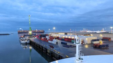 Yığınla renkli konteynır, rıhtımda büyük bir kargo gemisi ve akşam lojistiği sırasında suya yansıyan parlayan liman ışıkları olan endüstriyel liman terminalinin gece görüşü