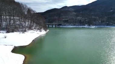 Buzların eridiği bahar başında sakin dağ gölü, kıyı şeridi boyunca eriyen karlar, ormanlık tepeler ve arka planda küçük bir köprü. Dağlardaki mevsimsel geçişi gösteren huzurlu doğal manzara