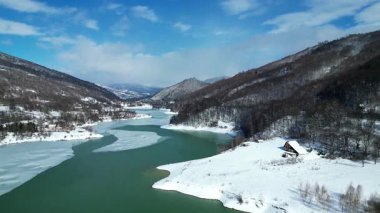 Baharın ilk zamanlarında eriyen buzlar, karlı tepeler, orman ve sahilde rahat bir ev ile eriyen bir dağ gölü manzarası. Huzurlu doğal manzara sakin bir dağ vadisinde mevsimsel geçiş gösteriyor.