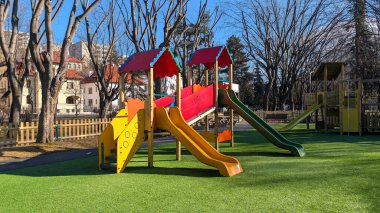 Güneşli bir günde, sakin bir şehir parkında yeşil yapay çimlerin üzerinde renkli slaytlarla ve ahşap yapılarla parlak ve güvenli çocuk parkı neşeli ve aile dostu bir atmosfer yaratıyor.