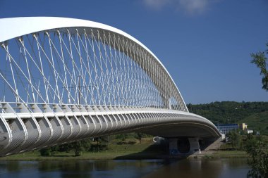 Modern beyaz kemerli bir köprü, yeşil tepeler ve parlak mavi bir gökyüzüne karşı güzel bir tasarım sergileyerek sakin bir nehri geçer. Sahne huzurlu doğayı yakalar..