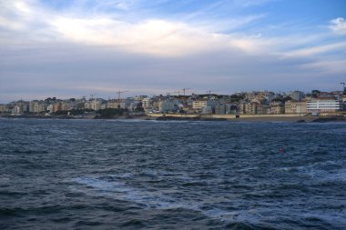 Sahilde sakin bir sahil kenti açılırken dalgalar yavaşça kıyıya çarpıyor. Uzun binalar ve turnalar gün batımında renkli bir gökyüzünün altında ufuk çizgisini işaretliyor..
