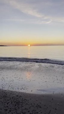 Gün batımındaki okyanus manzarası, yumuşak dalgaların kıyıya vurduğunu gösteriyor. Güneş ufukta batarken küçük bir yelkenli görünür..