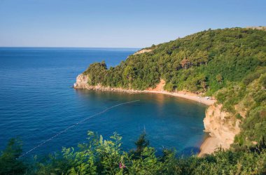 Budva yakınlarındaki Mogren plajı, ağaçlarla kaplı kayalık tepelerin arka planı, sakin mavi deniz. Mavi bulutsuz sabah gökyüzü. Karadağ ve Akdeniz