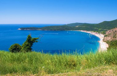 Budva yakınlarındaki Jaz sahilinde ağaçlarla kaplı kayalık tepelerin arka planında, sakin mavi deniz. Mavi bulutsuz sabah gökyüzü. Karadağ, Akdeniz 'de. Yüksek açı görünümü