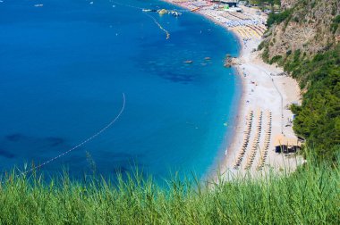 Budva yakınlarındaki Jaz sahilinde ağaçlarla kaplı kayalık tepelerin arka planında, sakin mavi deniz. Mavi bulutsuz sabah gökyüzü. Karadağ, Akdeniz 'de. Yüksek açı görünümü