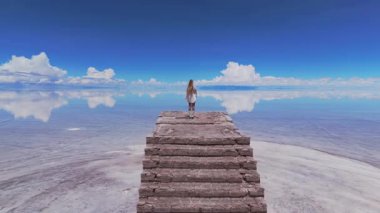 Bir kadın, Bolivya 'daki geniş Salar de Uyuni tuz düzlüklerinin ortasında, açık mavi gökyüzünün altında, tuğla bir yapının üzerinde duruyor..