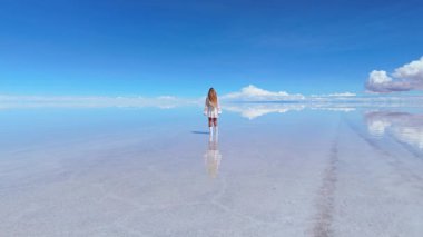 Bolivya 'daki geniş Uyuni Tuz Düzlükleri' nde yürüyen bir kadın, yansıması açık mavi bir gökyüzünün altındaki sığ suda yansıyor..