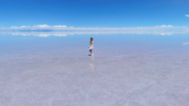 Bir kadın açık mavi gökyüzünün altında, Bolivya 'daki geniş Salar de Uyuni tuz düzlüklerinde yürüyor..