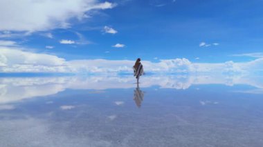 Bir kadın Bolivya 'da yansıtıcı bir tuzun üzerinde yürür, pançosu rüzgarda dalgalanır..