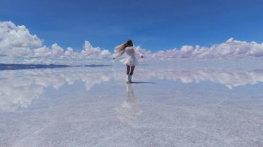Bir kadın zarif bir şekilde Bolivya 'daki geniş Salar de Uyuni tuz düzlüklerinde yürür, beyaz elbisesi parıldayan su yüzeyine karşıdır..