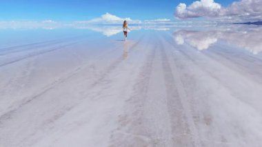 Bir kadın, Bolivya 'daki geniş Salar de Uyuni tuz düzlüklerinde yürür. Yansıması sığ sularda yankılanır..