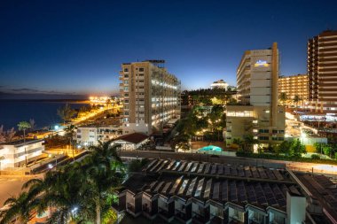 Kıyı otellerinin panoramik manzarası, palmiye ağaçları ve Gran Canaria 'da mavi saatte sakin okyanus. Parlak şehir ışıkları, koyu mavi gökyüzü ile tezat oluşturarak canlı bir seyahat ve tatil atmosferi yaratır..