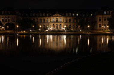 Stuttgart 'taki Yeni Saray gece aydınlandı, dramatik akşam bulutları altında sakin bir göle yansıdı. Çarpıcı bir dönüm noktası ve mimari bir gece manzarası.