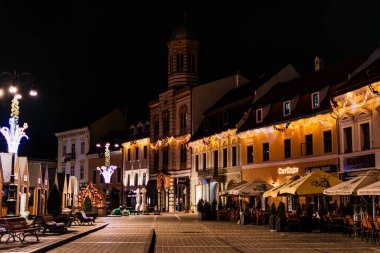 Brasov Konsey Meydanı 'ndaki tarihi binanın gece manzarası. Pencereler sıcak altın iplik ışıkları ve çelenklerle süslenmiş..