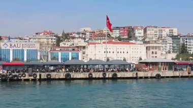 İstanbul 'daki Boğazın kıyısındaki restoranlarda dinlenen insanlar. Bahçeşehir Üniversitesi ve deniz kenarındaki kafelerin üzerinde Türk bayrağının dalgalandığı şehir manzarası.