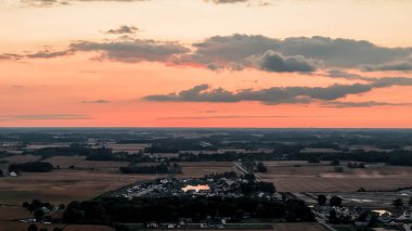 Gün batımında dramatik turuncu gökyüzü ve koyu bulutlu kırsal manzara. Akşam ışıklarıyla aydınlatılmış küçük kasaba ve tarım arazileri. Huzurlu bir atmosferi olan manzara manzarası.