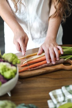 Tahta bir kesme tahtasıyla taze gökkuşağı havuçları dilimleyen bir kadın. Turuncu, sarı ve mor havuçlu renkli sebzeler, ön planda karnabahar. Sağlıklı beslenme, yemek hazırlığı, çiftlikten masaya konsept. Yüksek kalite fotoğraf
