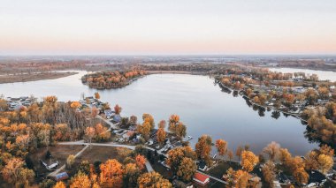 Canlı sonbahar ağaçlarıyla çevrili bir göl topluluğunun hava panoramik görüntüsü. Sakin su rengarenk sonbahar manzarasını yansıtıyor..