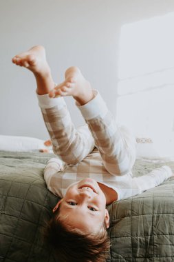 Mutlu çocuk rahat pijamalarıyla yatakta oynuyor, baş aşağı sabah ışığında gülümsüyor, evde tasasız bir çocukluk anı yaşıyor. Yüksek kalite fotoğraf