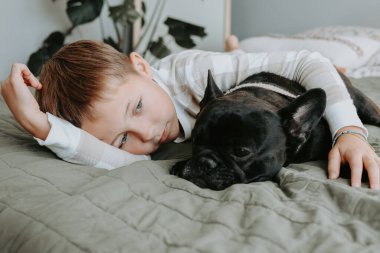 Genç bir çocuk, Fransız bulldoguyla yatakta dinleniyor. Evde sakin ve hassas bir anı paylaşıyor. Yüksek kalite fotoğraf