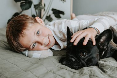 Yatakta bir Fransız Bulldog 'a sarılan gülümseyen genç bir çocuk, evde çocuklar ve evcil hayvanlarla rahat bir yaşam tarzı. Yüksek kalite fotoğraf
