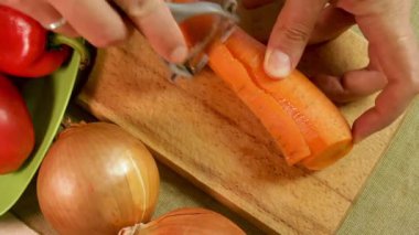 Erkek eller, soğan ve dolma biberlerle çevrili ahşap kesme tahtasıyla taze havuç soyarlar. Soyulmuş havuç dokusu ve tazeliği gösterir. Temel mutfak rutini ve sağlıklı yemek hazırlığı.