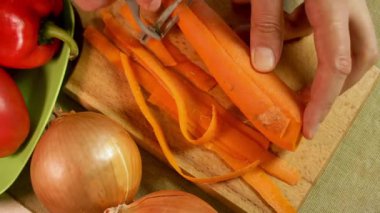 Adam havuç soyarken ahşap kabuk soyarken yanında soğan ve biberle tahta kesiyor. Soyulmuş şeritler tazeliği ön plana çıkarır. Rutin mutfak hazırlığı ve temiz yemek kavramı.