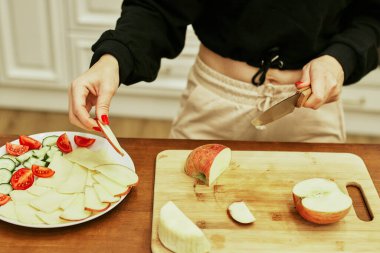 Genç kadın evdeki mutfakta sebze ve meyve kesiyor. Mutfak masasında taze meyve ve sebze var. Yemek pişirmek için malzemeler. Kapat.