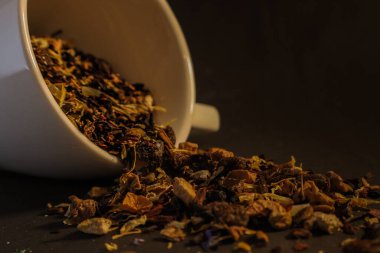 Beyaz seramik bir bardak, koyu bir yüzey boyunca kurumuş çay yaprakları ve otlardan oluşan bir karışım yayar. Canlı, renkli yapraklar etrafa saçılır ve topraksı, rahat bir atmosfer yaratır..