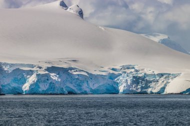 Antarktika Yarımadası 'nın kıyı şeridi karla kaplıydı. Mavi buz görünür. Önplanda okyanus, arkada bulutlu gökyüzü.