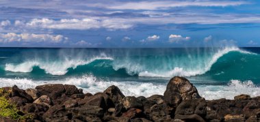 Hawaii 'nin Büyük Adası' nın batı sahilinde dalga kırılması. Köpük, ön planda volkanik kayalar. Mavi Pasifik Okyanusu, arka planda bulutlu gökyüzü.