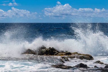 Hawaii 'nin büyük adasının doğu kıyısında dalga kırılması. Köpük, ön planda volkanik kayalar. Mavi Pasifik Okyanusu, arka planda bulutlu gökyüzü.
