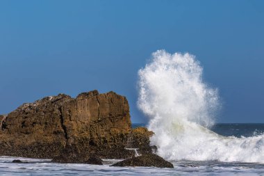 Malibu, California açıklarındaki büyük kayalara karşı dev dalgalar kırılıyor. Sprey havada yüksekte uçuyor, mavi gökyüzü uzakta..