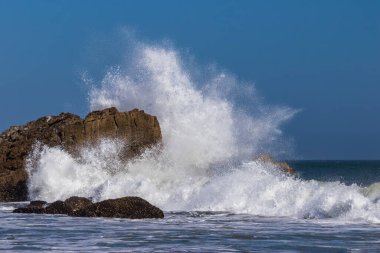 Malibu, California açıklarındaki büyük kayalara karşı dev dalgalar kırılıyor. Sprey havada yüksekte uçuyor, mavi gökyüzü uzakta..