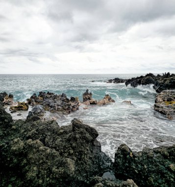 Karanlık volkanik kayalar, Pasifik Okyanusu 'nun çalkantılı dalgalarını kara kara düşünen bir gökyüzünün altında çerçeveler. Antik lav akıntıları tarafından şekillendirilen engebeli kıyı şeridi, huzursuz denizin yumuşak beyaz ve mavilerine karşıdır..
