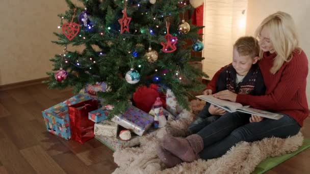 Feliz madre e hijo sentado bajo un árbol de Navidad y volteando álbum de fotos — Vídeo de stock