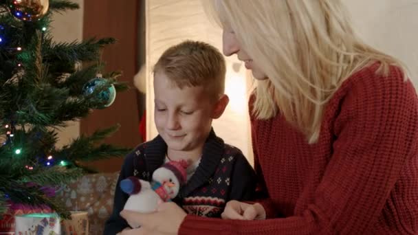 Hijo feliz con la madre sentada bajo el árbol de año nuevo y jugar con muñeco de nieve — Vídeos de Stock