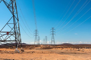 Ufuk çizgisine uzanan yüksek iletim kuleleri ve elektrik hatlarının olduğu çöl sahnesi. Güneşli gökyüzü ve kurak arazi, geniş ve izole bir arazi üzerinde altyapı, enerji iletimi ve uzak hizmet ağlarını vurguluyor..