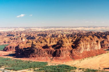 Katmanlı kırmızı kaya oluşumları, açık mavi gökyüzü ve tabanında yeşil bir iz olan çöl kanyonunu süpürüyor. Harrat 'ın bakış açısından bak. Seyahat, doğa ve açık hava macera temaları için mükemmel..