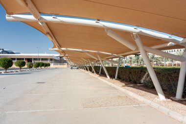 Geniş bir açık hava parkında beyaz direkler tarafından desteklenen büyük bej rengi yelkenler ve güneşli bir şehir kampüsünde modern, palmiye kaplı bir zemin yer alıyor..